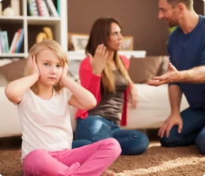 En ung jente sitter på gulvet og dekker for ørene mens foreldrene krangler i bakgrunnen. Foreldrene gestikulerer til hverandre, moren i rødt og faren i en blå skjorte.