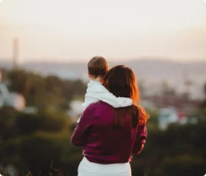 Mor som holder et lite barn med utsikt over en by ved solnedgang i Norge, som illustrerer familiestøtte og Barnetrygd-ytelser